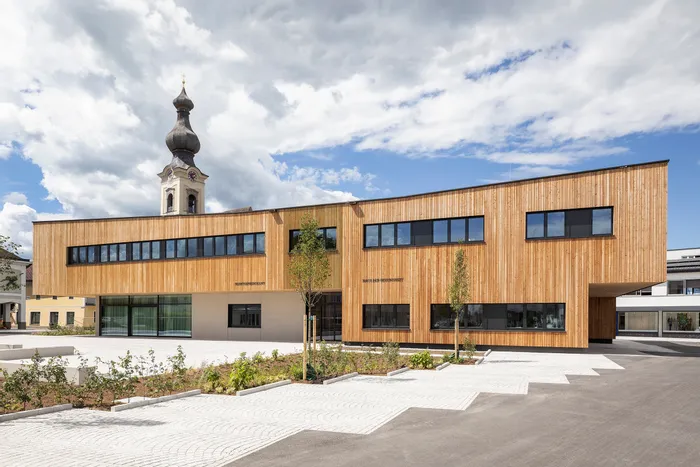 Modernes Schulgebäude mit Holzfassade neben barocker Kirchturmspitze unter bewölktem Himmel