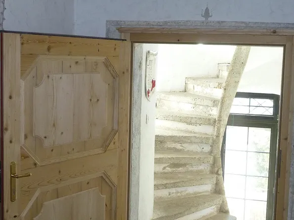 Geöffnete Holztür mit Blick auf eine steinerne Wendeltreppe und rundes Fenster darüber