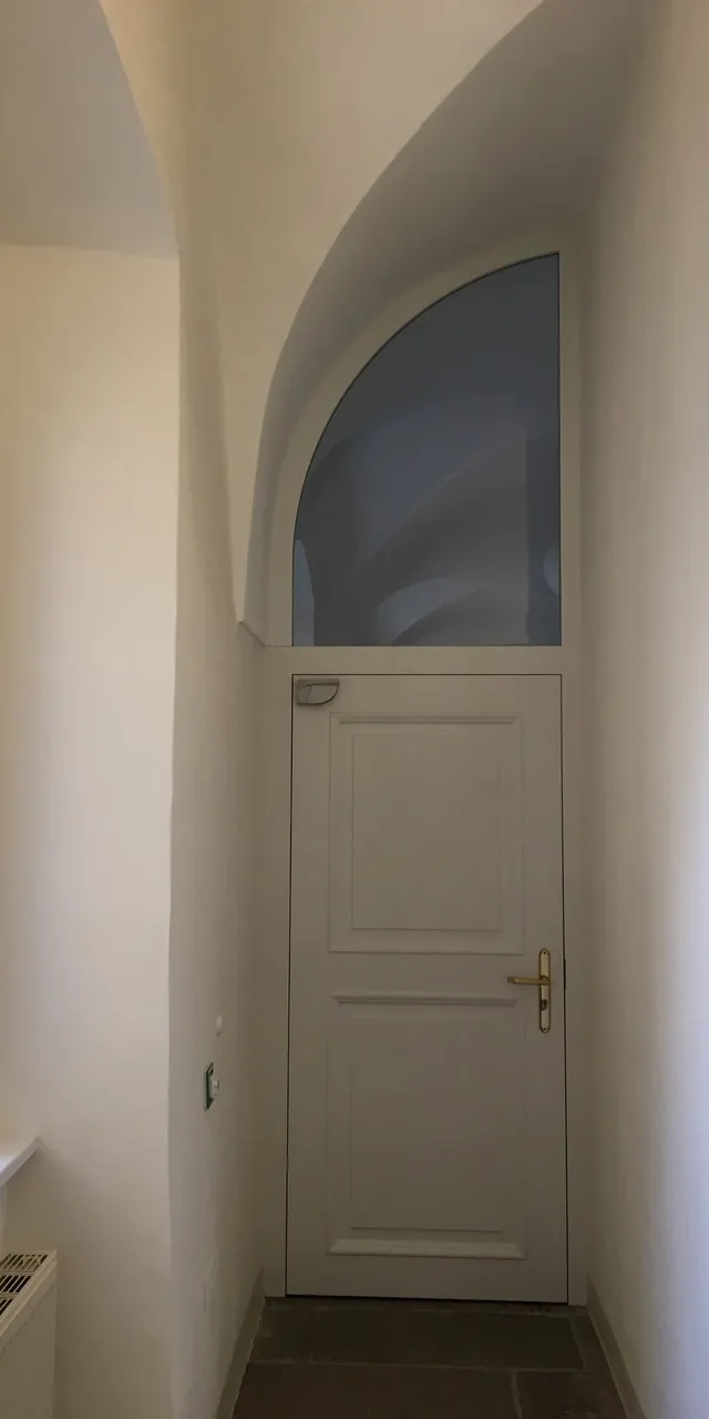 Weiße Holztür mit rundem Fenster oben in einem engen, hellen Flur mit Sicht auf ein Fenster