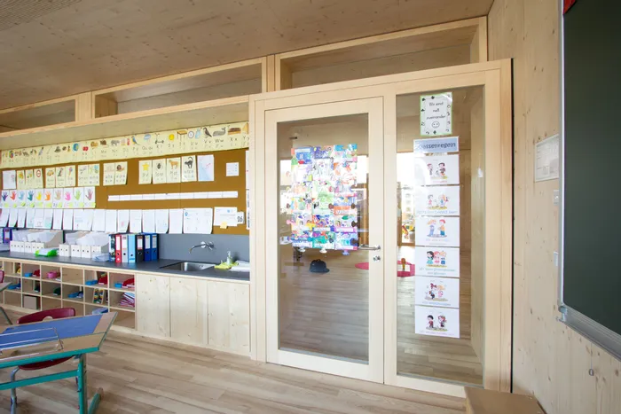 Heller Klassenzimmerraum mit Holzverkleidung, Kinderzeichnungen und Lernmaterialien an der Wand
