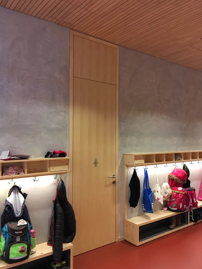 Kindergarderobe mit Holzbänken und Jacken vor einer hohen Tür mit Mädchensymbol