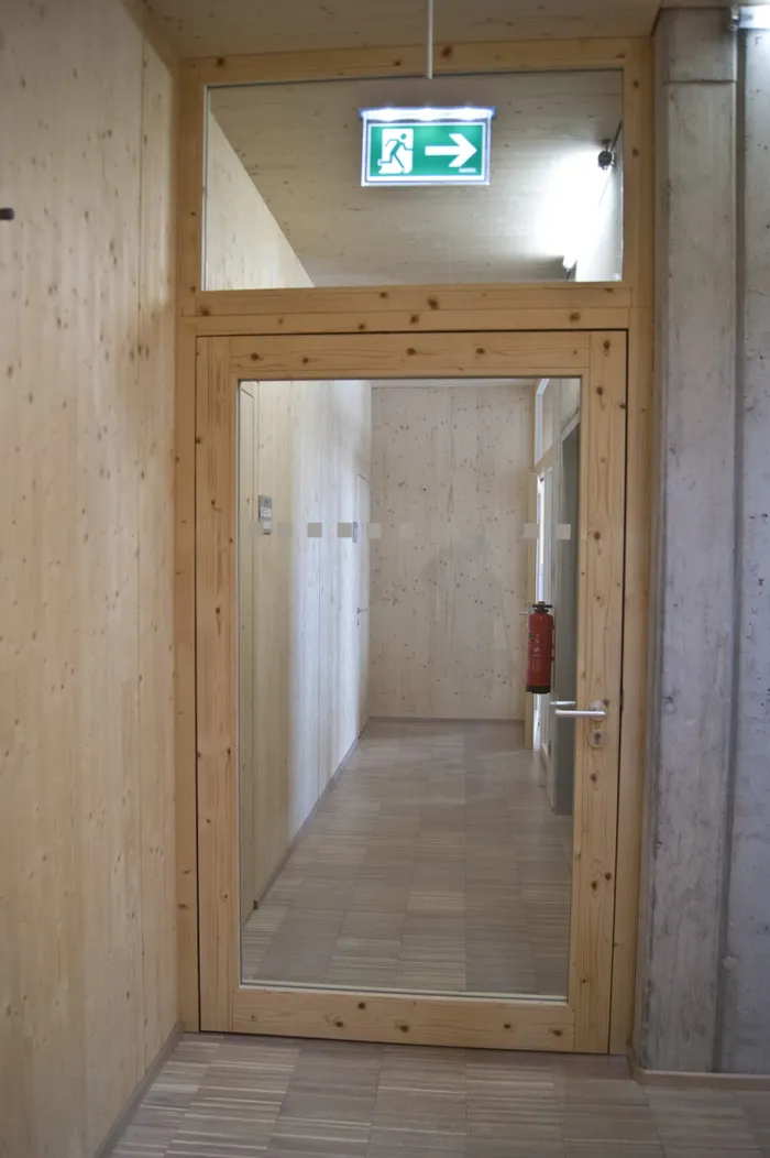 Flur mit Holzwänden, Glastür und grünem Notausgangsschild mit Pfeil nach rechts
