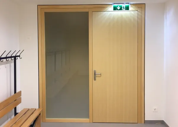 Umkleideraum mit Holzbank, Kleiderhaken und Tür mit grünem Notausgangsschild