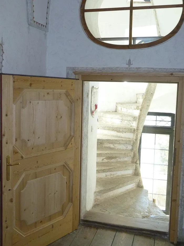 Geöffnete Holztür mit Blick auf eine steinerne Wendeltreppe und rundes Fenster darüber