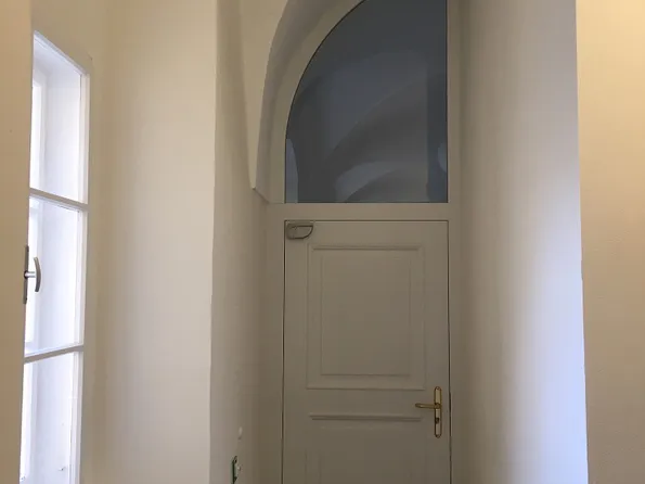 Weiße Holztür mit rundem Fenster oben in einem engen, hellen Flur mit Sicht auf ein Fenster