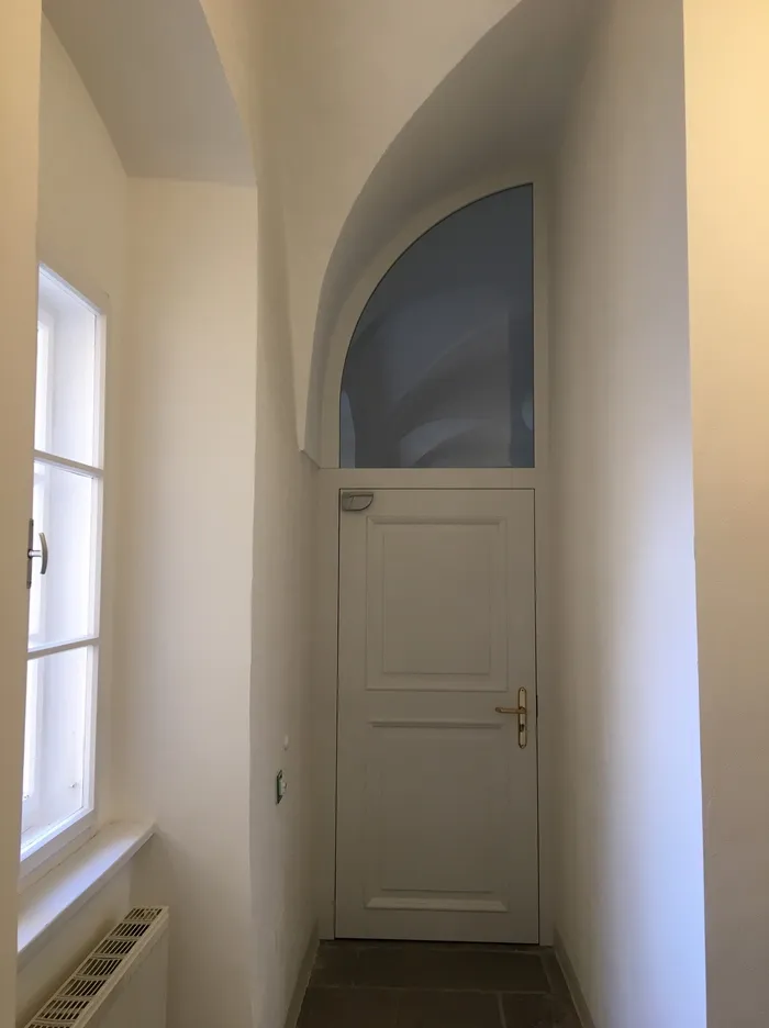 Weiße Holztür mit rundem Fenster oben in einem engen, hellen Flur mit Sicht auf ein Fenster