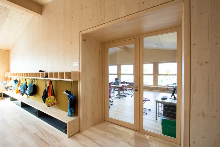 Holzverkleideter Schulflur mit Garderobe und Blick in ein Klassenzimmer mit Tischen und Stühlen