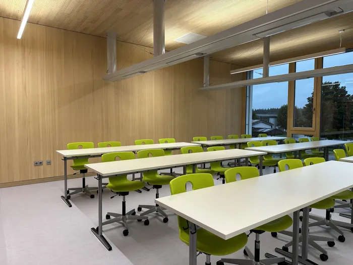 Moderner Klassenraum mit weißen Tischen und grünen Stühlen vor Holzverkleidung und großen Fenstern