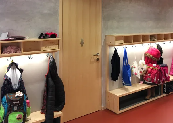 Kindergarderobe mit Holzbänken und Jacken vor einer hohen Tür mit Mädchensymbol