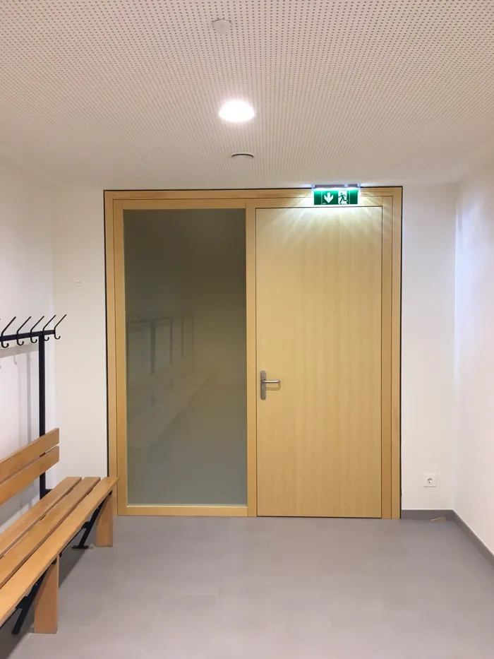 Umkleideraum mit Holzbank, Kleiderhaken und Tür mit grünem Notausgangsschild