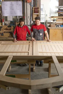 Zwei Schreiner begutachten eine große Holztür in einer Werkstatt mit Holzbauplänen an der Wand