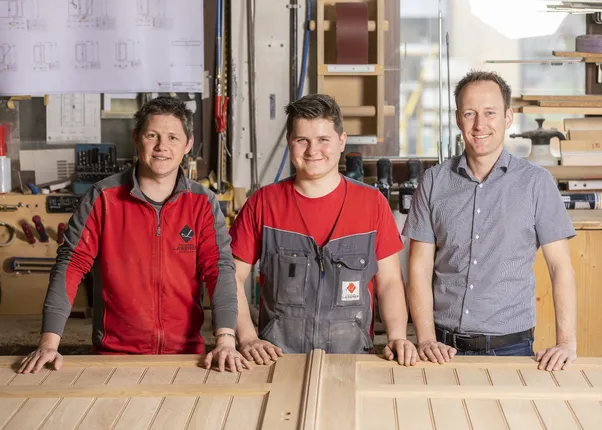 Drei Schreiner stehen lächelnd in einer Werkstatt hinter einer Holztür