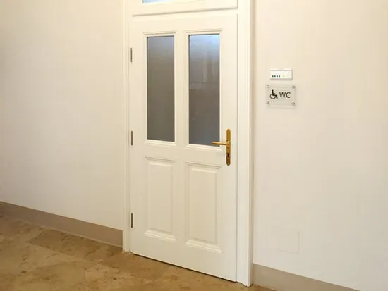 Weiße WC-Tür mit Glaseinsätzen und Schild für barrierefreie Toilette an der Wand