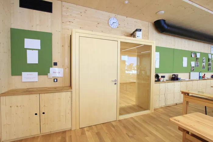 Moderner Klassenraum mit Holzmöbeln, Glastür, grünen Pinnwänden und Wanduhren