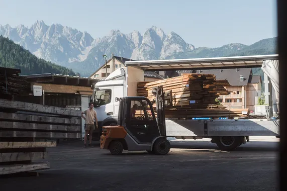 Gabelstapler lädt Holzbretter von LKW in einem Sägewerk mit Alpen im Hintergrund